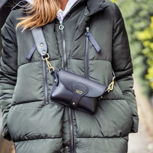 Puede incluir: Un bolso bandolera de cuero negro con una cadena dorada y las iniciales "MD" grabadas en la parte delantera. El bolso se lleva sobre una chaqueta acolchada verde.