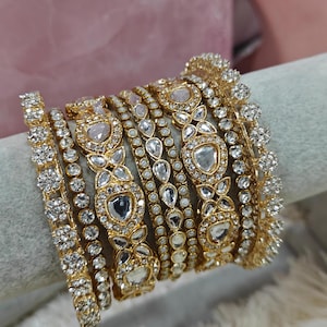 Peut inclure: Un ensemble de bracelets dorés avec des pierres blanches et des accents de perles. Les bracelets sont empilés les uns sur les autres et présentent un design décoratif.