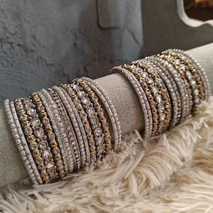 Peut inclure: Un ensemble de bracelets blancs et dorés avec des strass et des perles. Les bracelets sont empilés sur une surface blanche et moelleuse.