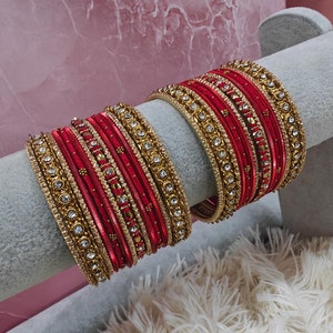 Op de afbeelding: Een set rode en gouden armbanden met strass. De armbanden zijn gestapeld op een witte displaystandaard.