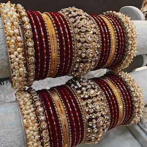 May include: A set of maroon and gold bangles with white pearl accents. The bangles are decorated with intricate designs and rhinestones.