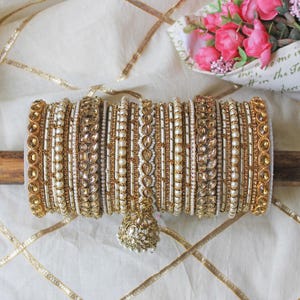 May include: A set of gold and white bangles with pearl and gemstone embellishments. The bangles are stacked on a wooden surface and have a decorative charm hanging from the bottom.