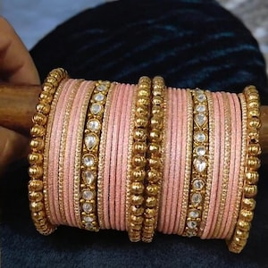 May include: A stack of pink and gold bangles with clear rhinestones. The bangles are made of a thin, flexible material and are decorated with small gold beads and rhinestones.
