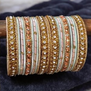 Könnte beinhalten: Eine Sammlung goldfarbener Armreifen mit aufwendigen Details. Die Armreifen weisen abwechselnd goldene, rosa und hellgrüne Bänder auf, die mit glitzernden Verzierungen versehen sind. Der Schmuck wird auf einem Holzstab vor dunklem Hintergrund präsentiert.