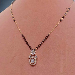 Puede incluir: Un collar dorado con un colgante con piedras preciosas transparentes. El collar tiene una cadena delicada y est&aacute; adornado con peque&ntilde;as cuentas oscuras. El colgante tiene forma de l&aacute;grima con un grupo de piedras brillantes.