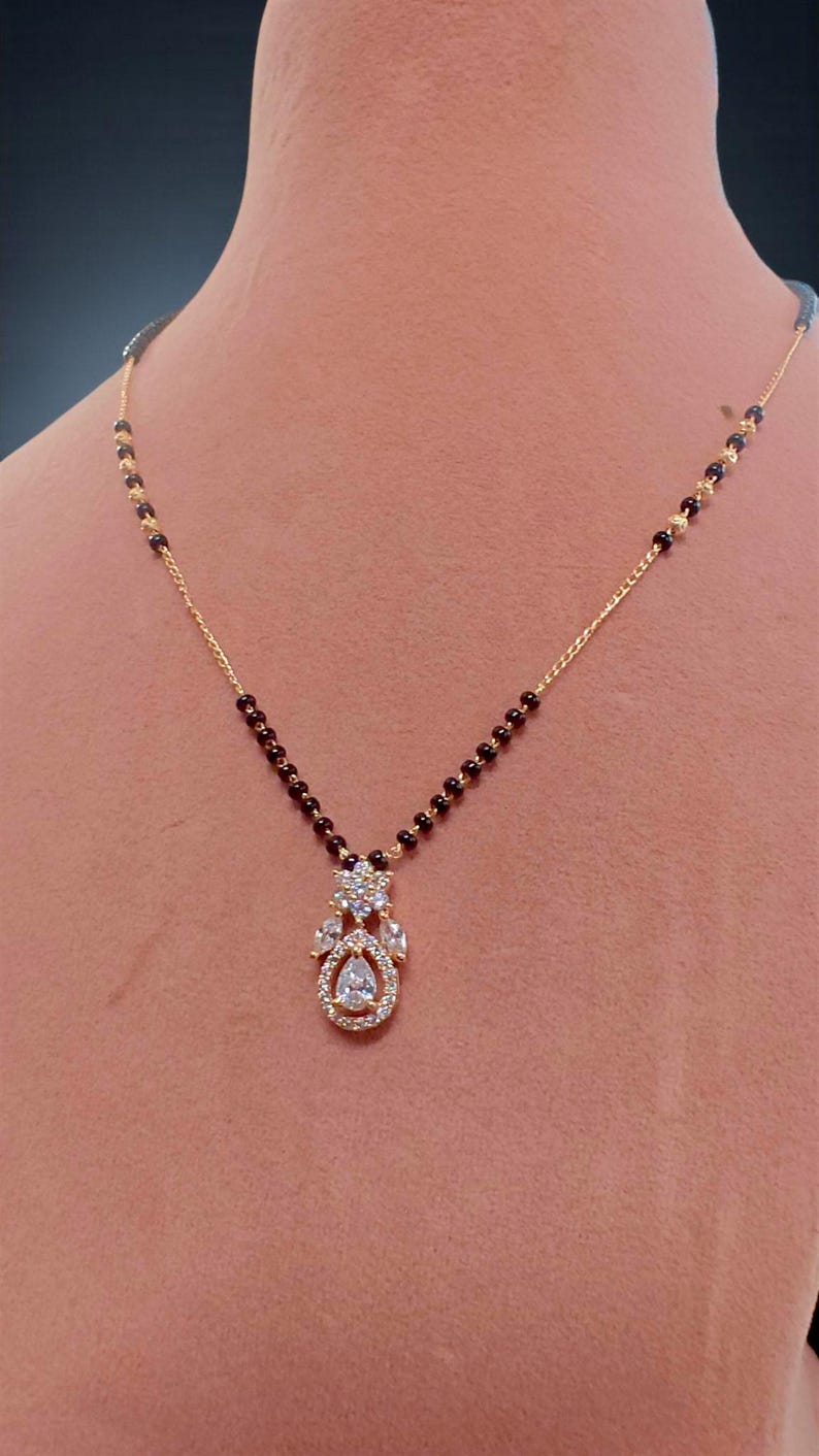 Puede incluir: Un collar dorado con un colgante con piedras preciosas transparentes. El collar tiene una cadena delicada y est&aacute; adornado con peque&ntilde;as cuentas oscuras. El colgante tiene forma de l&aacute;grima con un grupo de piedras brillantes.