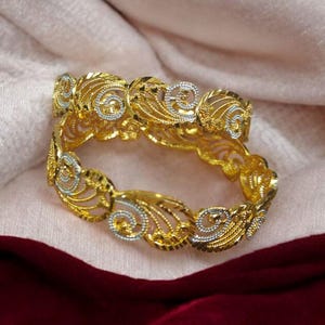 Puede incluir: Dos brazaletes dorados con intrincados dise&ntilde;os calados. Los brazaletes presentan una mezcla de acentos dorados y plateados, con un patr&oacute;n floral y arremolinado. Las joyas se exhiben sobre una tela rosa y burdeos.