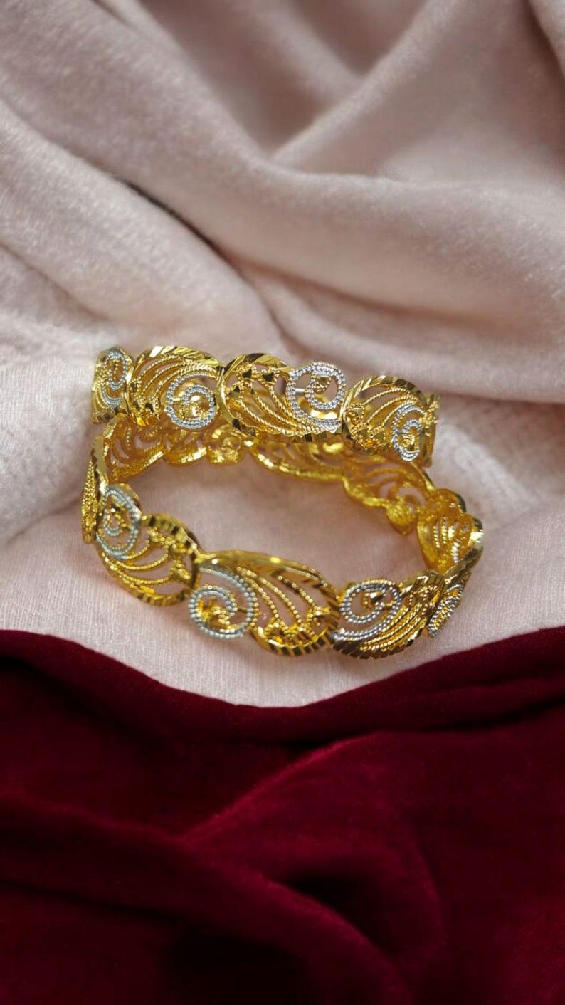 Puede incluir: Dos brazaletes dorados con intrincados dise&ntilde;os calados. Los brazaletes presentan una mezcla de acentos dorados y plateados, con un patr&oacute;n floral y arremolinado. Las joyas se exhiben sobre una tela rosa y burdeos.