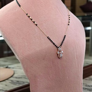 Puede incluir: Un collar dorado con un colgante con piedras preciosas transparentes y una cadena de cuentas negras. El collar se exhibe en un maniqu&iacute; rosa, mostrando su dise&ntilde;o. El colgante tiene forma de l&aacute;grima.