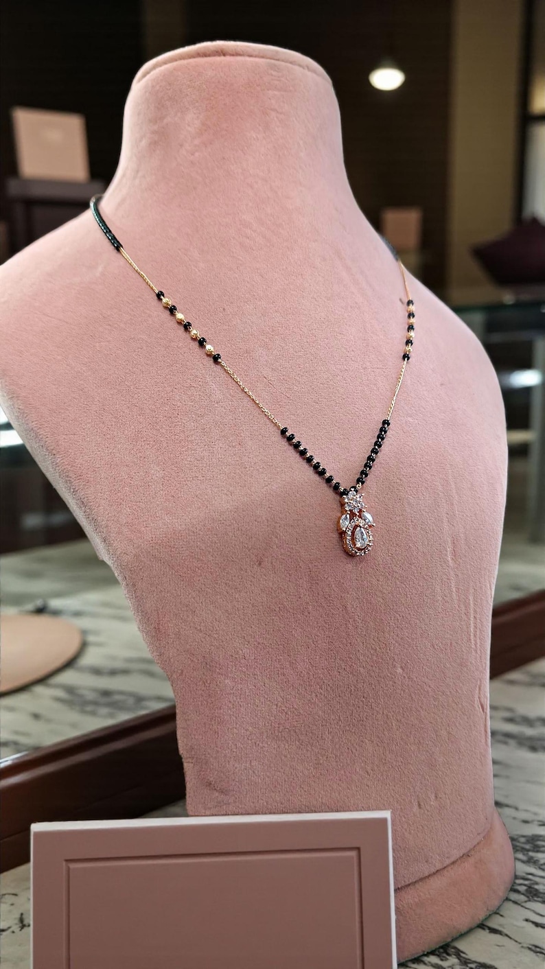 Puede incluir: Un collar dorado con un colgante con piedras preciosas transparentes y una cadena de cuentas negras. El collar se exhibe en un maniqu&iacute; rosa, mostrando su dise&ntilde;o. El colgante tiene forma de l&aacute;grima.