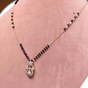 Puede incluir: Un collar dorado con un colgante con piedras preciosas transparentes y una piedra central en forma de l&aacute;grima. La cadena tiene cuentas negras. El collar se exhibe sobre una superficie rosa.