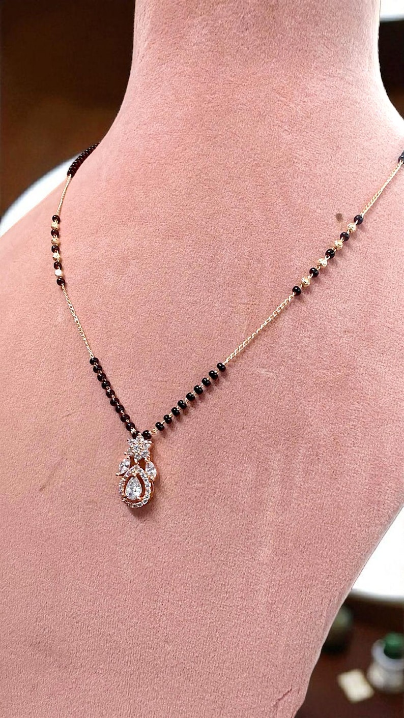 Puede incluir: Un collar dorado con un colgante con piedras preciosas transparentes y una piedra central en forma de l&aacute;grima. La cadena tiene cuentas negras. El collar se exhibe sobre una superficie rosa.