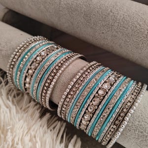 Könnte beinhalten: Ein Set aus silbernen und türkisfarbenen Armreifen mit Strasssteinen. Die Armreifen sind übereinander gestapelt und mit kleinen, funkelnden Steinen verziert.