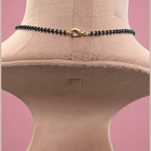 Puede incluir: Un collar de cuentas negras con un cierre dorado se exhibe en un maniqu&iacute;. El collar est&aacute; compuesto por peque&ntilde;as cuentas negras redondas. El cierre tiene un dise&ntilde;o simple y curvo. El maniqu&iacute; es de color beige claro, sobre un fondo rosa.