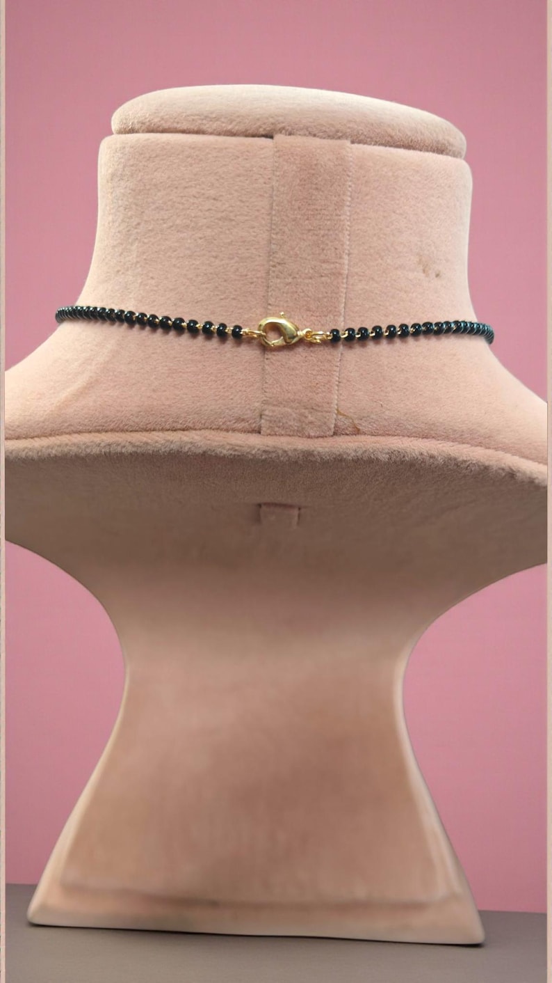 Puede incluir: Un collar de cuentas negras con un cierre dorado se exhibe en un maniqu&iacute;. El collar est&aacute; compuesto por peque&ntilde;as cuentas negras redondas. El cierre tiene un dise&ntilde;o simple y curvo. El maniqu&iacute; es de color beige claro, sobre un fondo rosa.