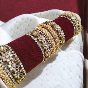 Op de afbeelding: Een set gouden armbanden met gespiegelde en parel accenten. De armbanden zijn gestapeld op een bordeauxrode fluwelen basis.