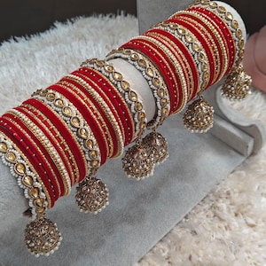 May include: A set of red and gold bangles with white beads and decorative charms. The bangles are stacked on a gray surface.