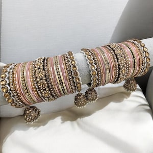 May include: A set of pink and gold bangles with pearl and rhinestone embellishments. The bangles are stacked on a white surface and have decorative charms.