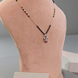 Puede incluir: Un collar dorado con una cadena de cuentas negras y un colgante con piedras preciosas transparentes. El collar se exhibe en un maniqu&iacute; rosa, mostrando su dise&ntilde;o y detalles. El colgante tiene un dise&ntilde;o floral.