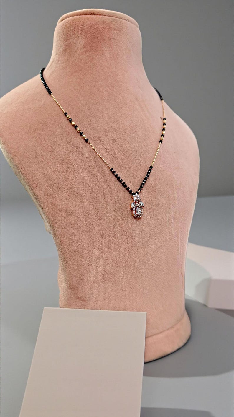 Puede incluir: Un collar dorado con una cadena de cuentas negras y un colgante con piedras preciosas transparentes. El collar se exhibe en un maniqu&iacute; rosa, mostrando su dise&ntilde;o y detalles. El colgante tiene un dise&ntilde;o floral.