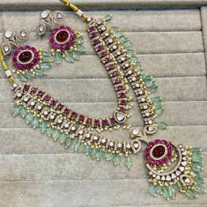 Peut inclure: Un collier à deux rangs avec un grand pendentif orné d'une pierre rose entourée de petites pierres blanches. Le collier est orné de perles vertes et blanches et de pierres roses. Des boucles d'oreilles assorties sont également incluses.