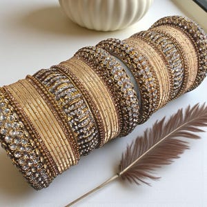 May include: A set of gold-coloured bangles with detailed patterns and clear, sparkling stones. The bangles are stacked, displaying different textures and designs. A brown feather lies beside them, adding a touch of elegance.