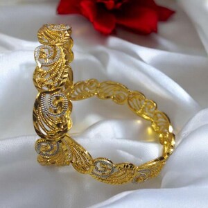 Puede incluir: Dos pulseras doradas con intrincados dise&ntilde;os de filigrana y volutas. Las pulseras est&aacute;n adornadas con detalles plateados y descansan sobre una superficie de sat&eacute;n blanco, con una flor roja en el fondo. La joyer&iacute;a es ornamentada y elegante.