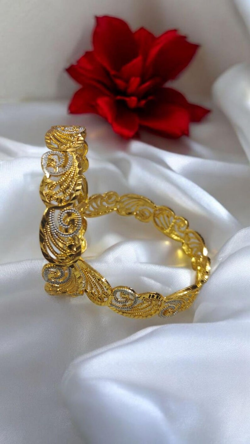Puede incluir: Dos pulseras doradas con intrincados dise&ntilde;os de filigrana y volutas. Las pulseras est&aacute;n adornadas con detalles plateados y descansan sobre una superficie de sat&eacute;n blanco, con una flor roja en el fondo. La joyer&iacute;a es ornamentada y elegante.