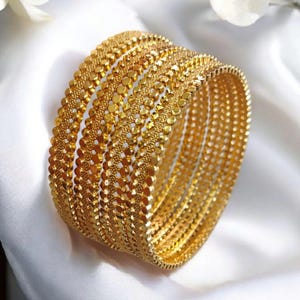 Puede incluir: Un conjunto de pulseras doradas con un diseño texturizado y punteado. Las pulseras están dispuestas sobre una superficie blanca y sedosa, con algunas flores blancas en el fondo. Las pulseras son de un material dorado brillante.