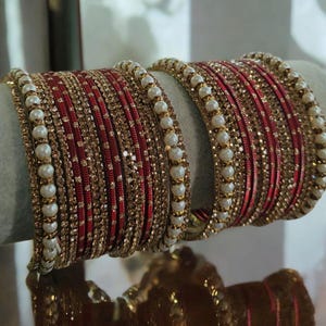 Pode incluir: Um par de pulseiras vermelhas e douradas com detalhes de pérolas brancas. As pulseiras são empilhadas, exibindo um design detalhado com enfeites brilhantes. As pérolas são espaçadas uniformemente.