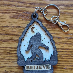 May include: A black wooden keychain with a silhouette of Bigfoot walking in front of a crescent moon and stars. The keychain has the word "BELIEVE" engraved on the bottom.