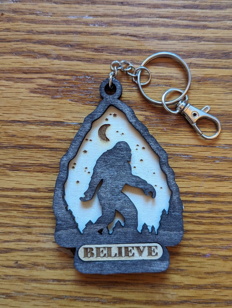 May include: A black wooden keychain with a silhouette of Bigfoot walking in front of a crescent moon and stars. The keychain has the word "BELIEVE" engraved on the bottom.