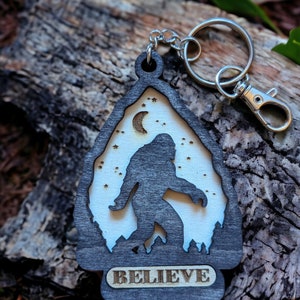 May include: A wooden keychain with a silhouette of Bigfoot walking in front of a crescent moon and stars. The keychain is black with a white background and the word "BELIEVE" is engraved on the bottom.