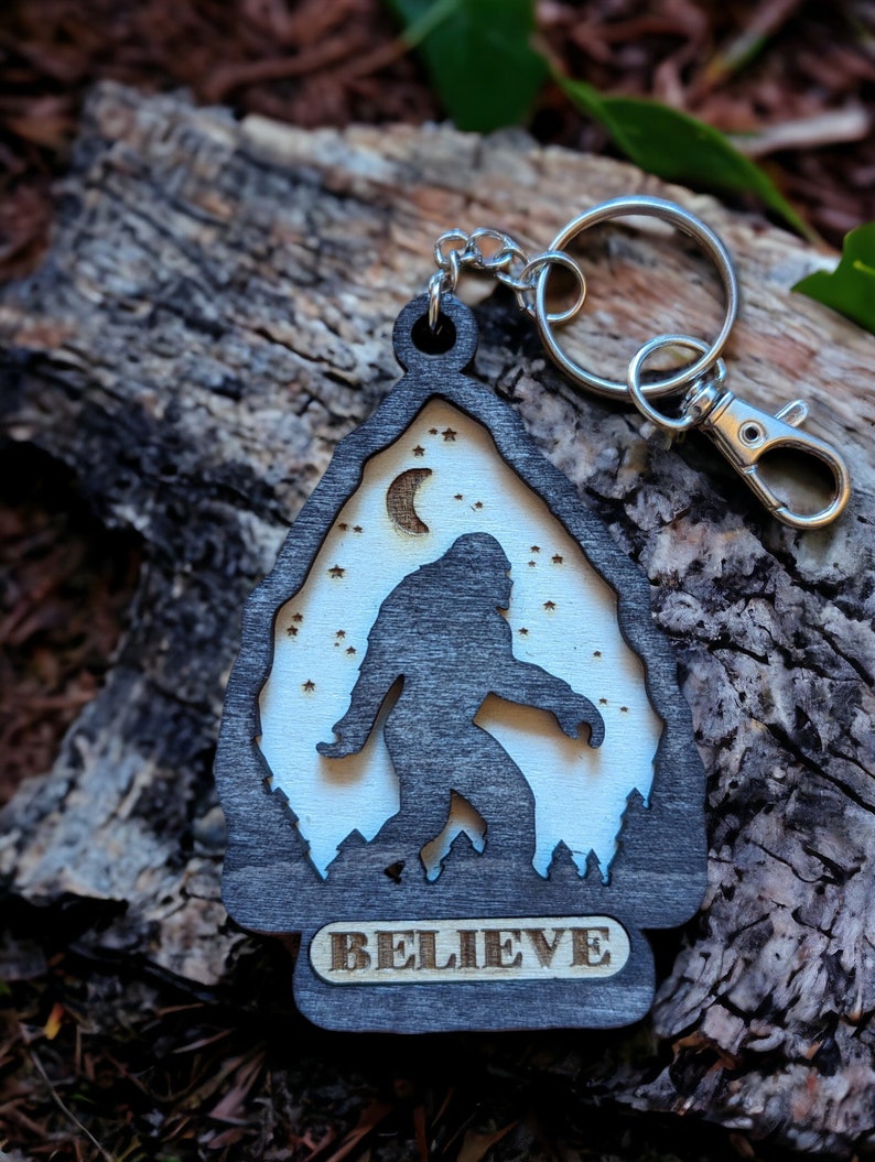 May include: A wooden keychain with a silhouette of Bigfoot walking in front of a crescent moon and stars. The keychain is black with a white background and the word "BELIEVE" is engraved on the bottom.