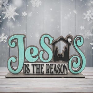 May include: A wooden Christmas decoration with the words "Jesus is the Reason" in teal and gray. The design includes a silhouette of the nativity scene and snowflake accents, perfect for holiday decor.