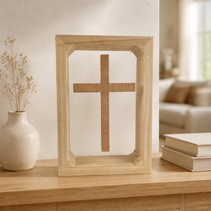 May include: A light-colored wooden rectangular frame containing a suspended, centered cross. The cross features a textured, linear grain pattern and is held within the open space of the frame, which has beveled inner edges and a smooth finish.