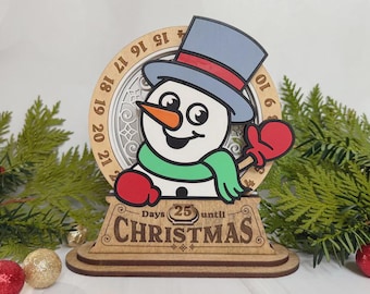Puede incluir: Decoración de cuenta regresiva navideña de madera con un muñeco de nieve con sombrero azul, nariz naranja y guantes rojos. El diseño circular tiene números alrededor del borde y el texto "Days 25 until CHRISTMAS" en la base.