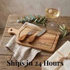 Può includere: Un tagliere in legno con manico, una forchetta e un coltello, tutti con manici in legno e la lettera "S" incisa. La tavola è incisa con il nome "Stephanie". È presente anche un bicchiere di champagne con un fiore.
