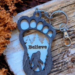 May include: A wooden keychain shaped like a Bigfoot footprint with a silhouette of Bigfoot inside. The keychain has the word "Believe" written on it and is attached to a silver key ring.
