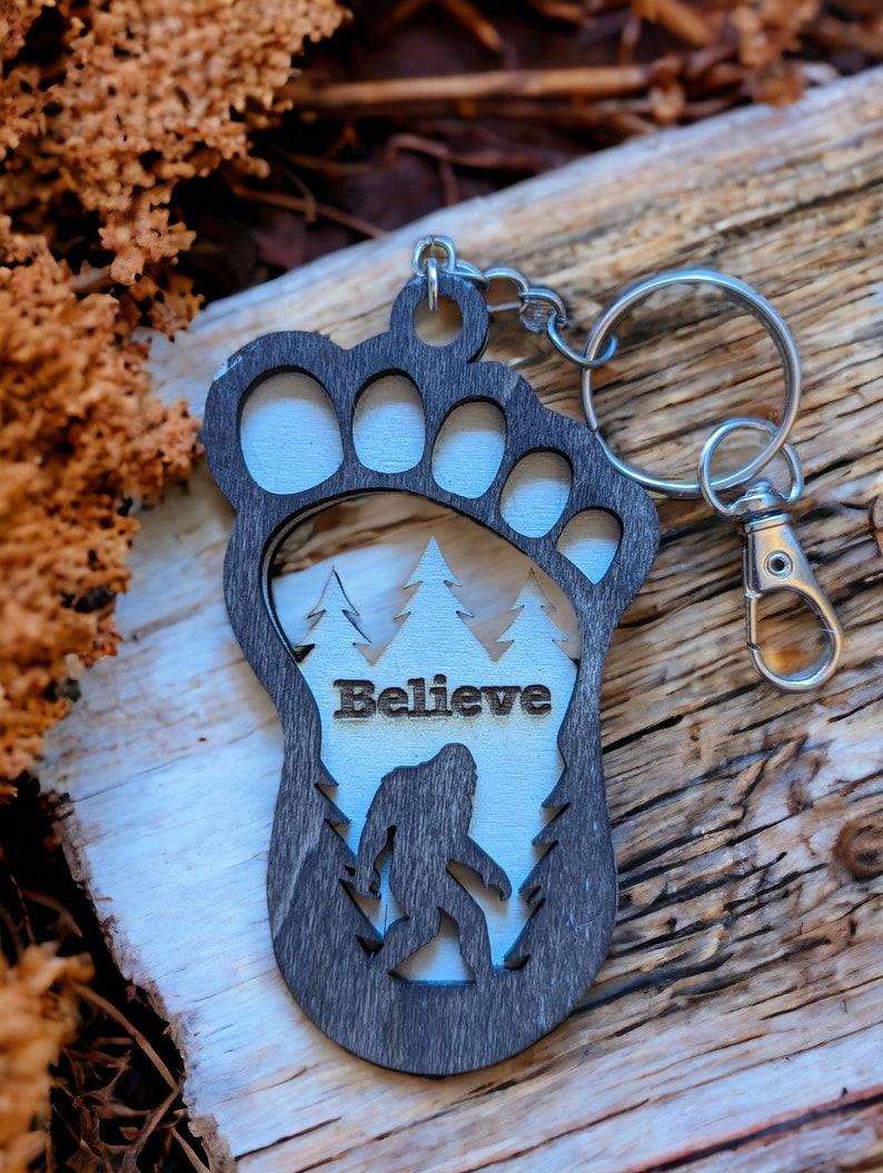 May include: A wooden keychain shaped like a Bigfoot footprint with a silhouette of Bigfoot inside. The keychain has the word "Believe" written on it and is attached to a silver key ring.
