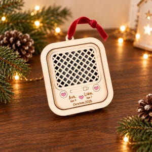 May include: A square wooden ornament shaped like a vintage radio with a red velvet ribbon hanger. The front features a black lattice speaker grille, two pink heart icons, and engraved text reading Ava, Age 4, Liam, Age 7, and Christmas 2026.