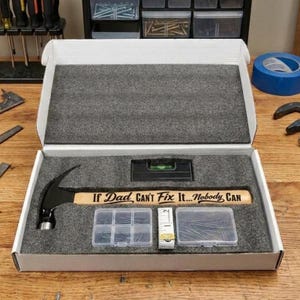 May include: A white cardboard gift box with grey foam inserts containing a black claw hammer with a wooden handle engraved with the text If Dad Can't Fix It... Nobody Can, a small black level, a clear plastic organizer with nails, and a measuring tape.