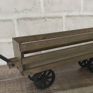 Könnte beinhalten: Ein kleiner Holzkarren mit langem Griff und vier schwarzen Rädern. Die Ladefläche des Wagens ist rechteckig und besteht aus horizontalen Holzlatten. Der Griff ist schwarz und an der Vorderseite des Wagens befestigt.