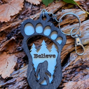 May include: A black wooden keychain shaped like a Bigfoot footprint with a white silhouette of Bigfoot walking through a forest inside the footprint. The keychain has the word "Believe" written above the Bigfoot silhouette.