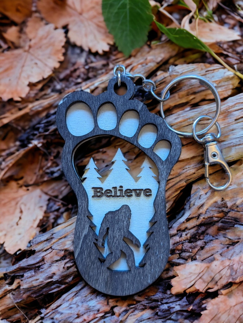 May include: A black wooden keychain shaped like a Bigfoot footprint with a white silhouette of Bigfoot walking through a forest inside the footprint. The keychain has the word "Believe" written above the Bigfoot silhouette.