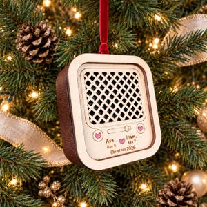 May include: A square wooden Christmas ornament shaped like a vintage radio with a dark brown side and a light wood front. The front features a grid speaker pattern, two small pink heart icons, and text reading Ava, Age 4, Liam, Age 7, Christmas 2026.