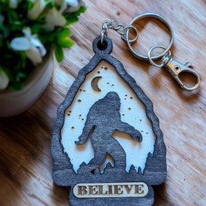 May include: A wooden keychain shaped like a mountain with a silhouette of Bigfoot walking in front of a crescent moon and stars. The keychain has the word "BELIEVE" engraved on the bottom.