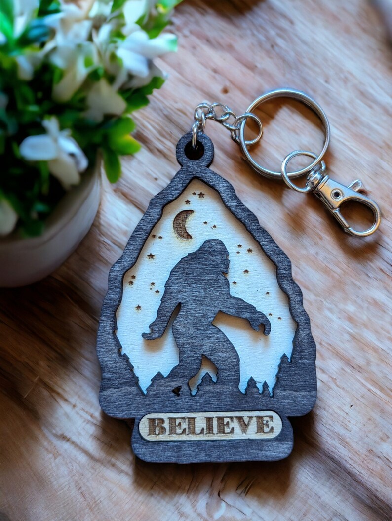 May include: A wooden keychain shaped like a mountain with a silhouette of Bigfoot walking in front of a crescent moon and stars. The keychain has the word "BELIEVE" engraved on the bottom.