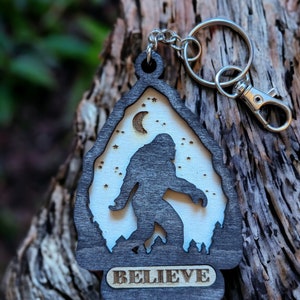 May include: A wooden keychain with a silhouette of Bigfoot walking in front of a crescent moon and stars. The keychain is dark brown with a light brown background and the word "BELIEVE" is engraved on the bottom.