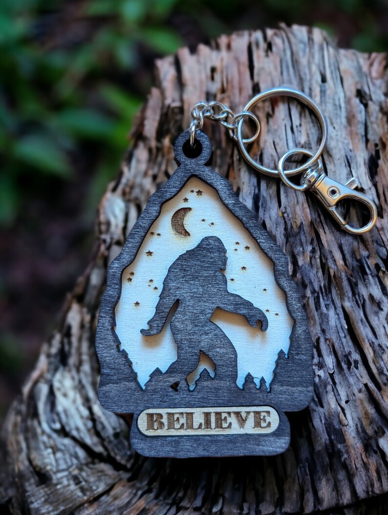 May include: A wooden keychain with a silhouette of Bigfoot walking in front of a crescent moon and stars. The keychain is dark brown with a light brown background and the word "BELIEVE" is engraved on the bottom.